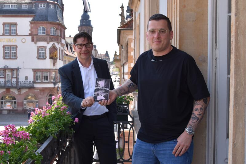 Auto Daniel Gebhart (rechts) &uuml;berreicht Oberb&uuml;rgermeister Thomas Hirsch ein Exemplar seines Buchs &raquo;Strassenstaub&laquo;. 